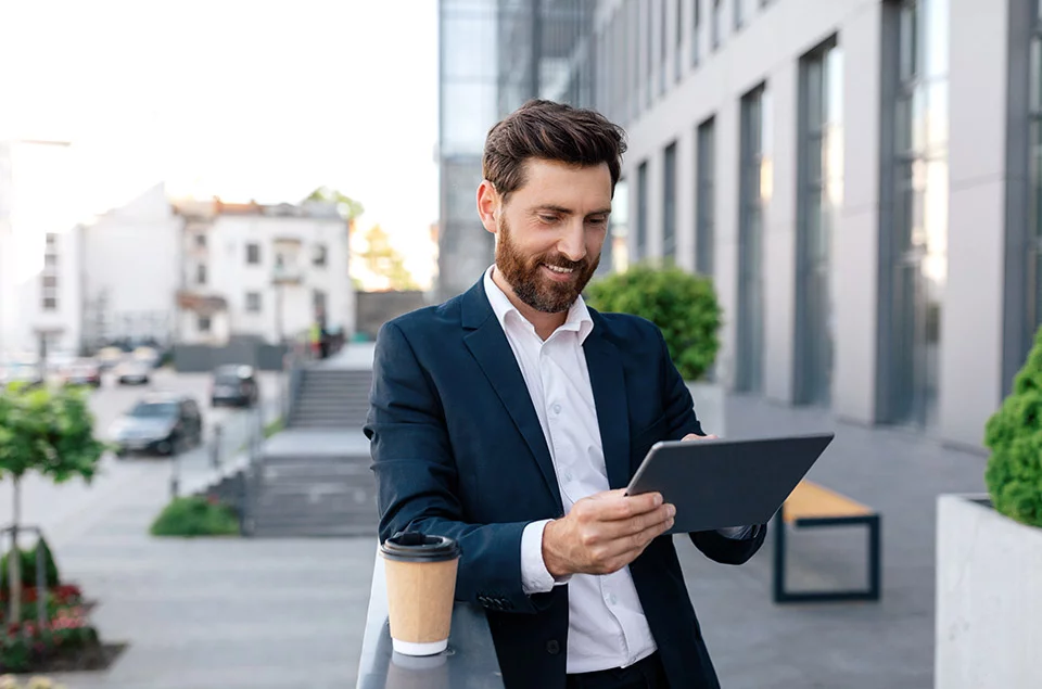 Mann steht mit einem Kaffee und seinem Ipad auf einem Balkon